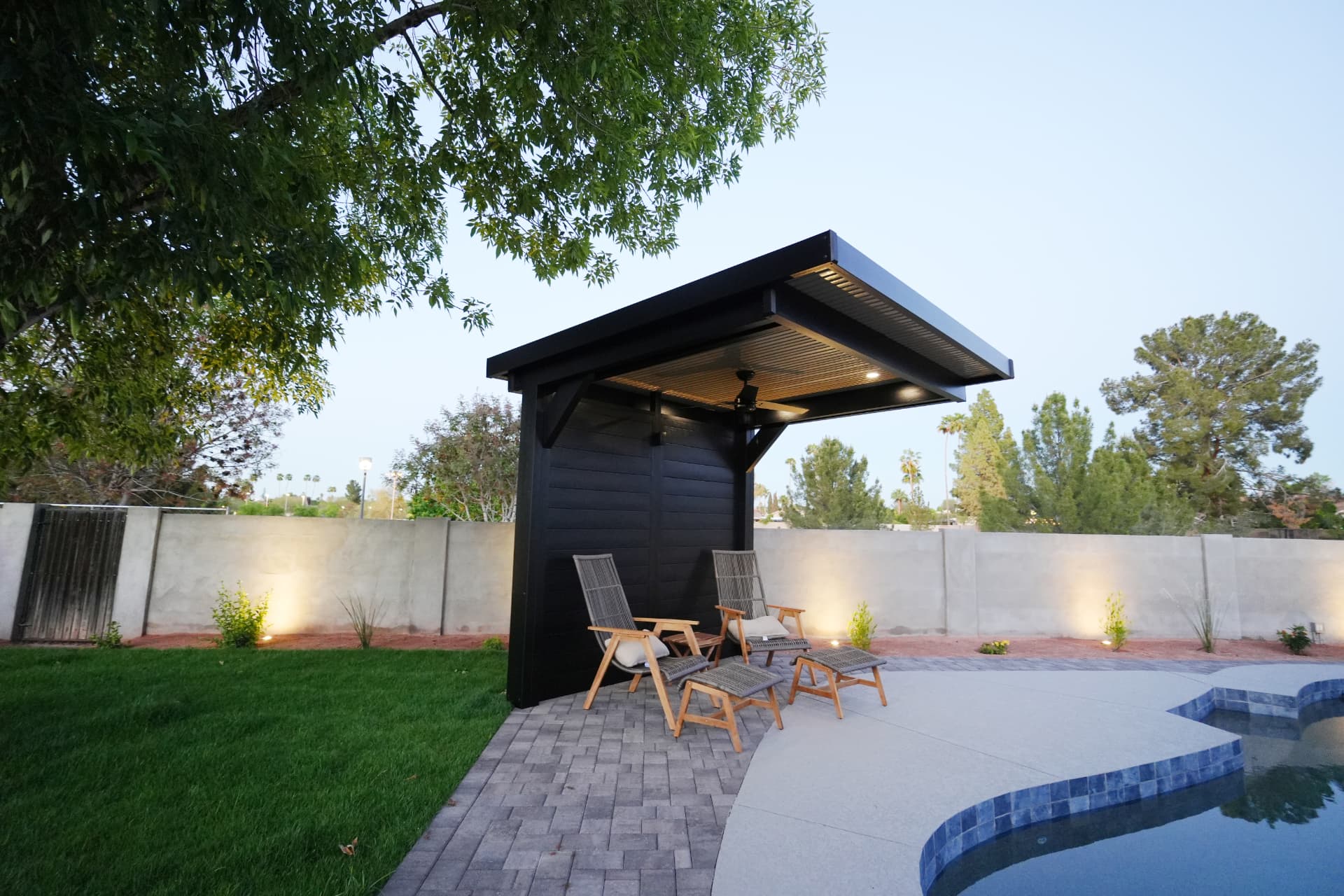 Black louvered cabana with lounge chairs beside a pool at dusk