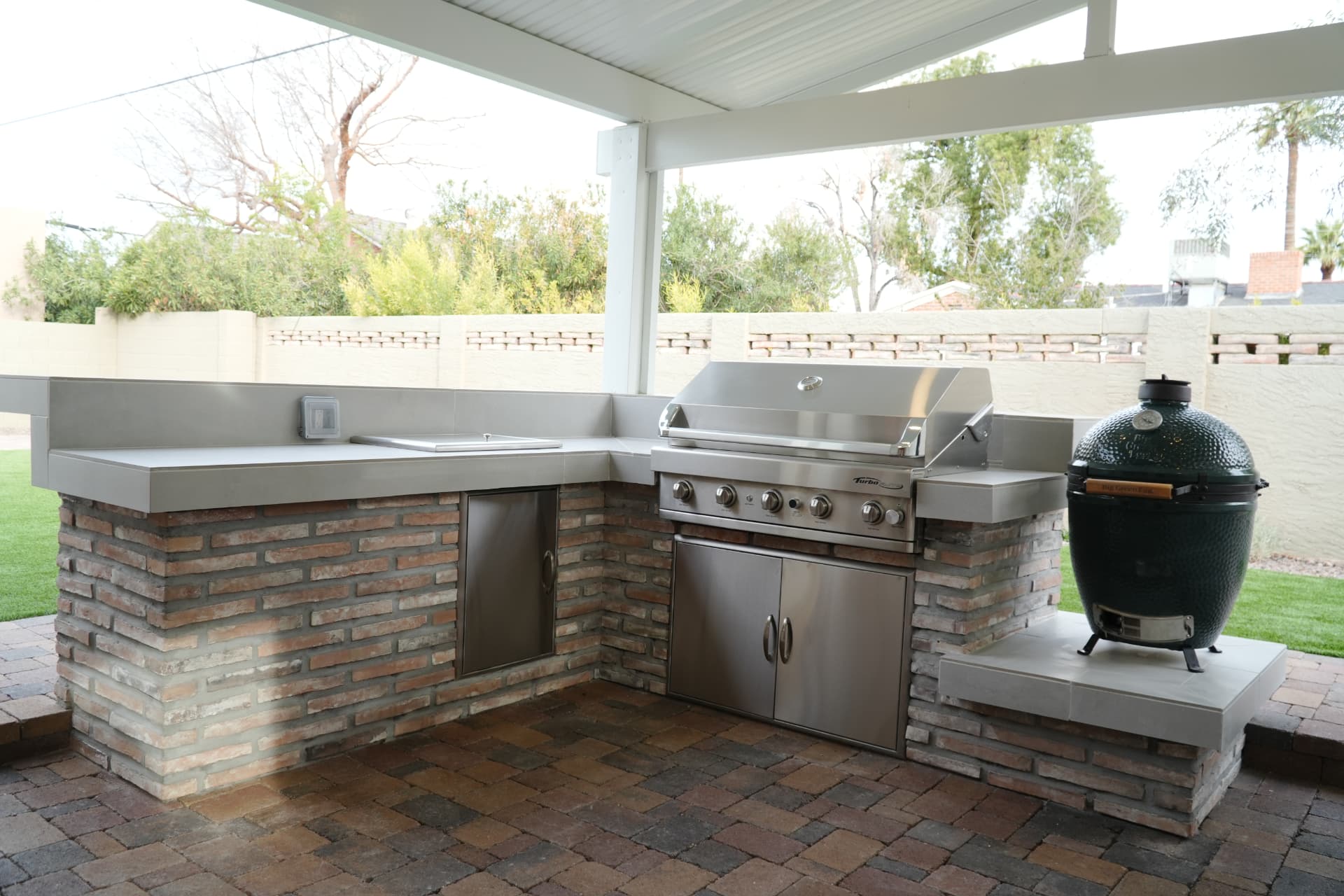 L-shaped brick outdoor kitchen with stainless grill, sink and kamado smoker