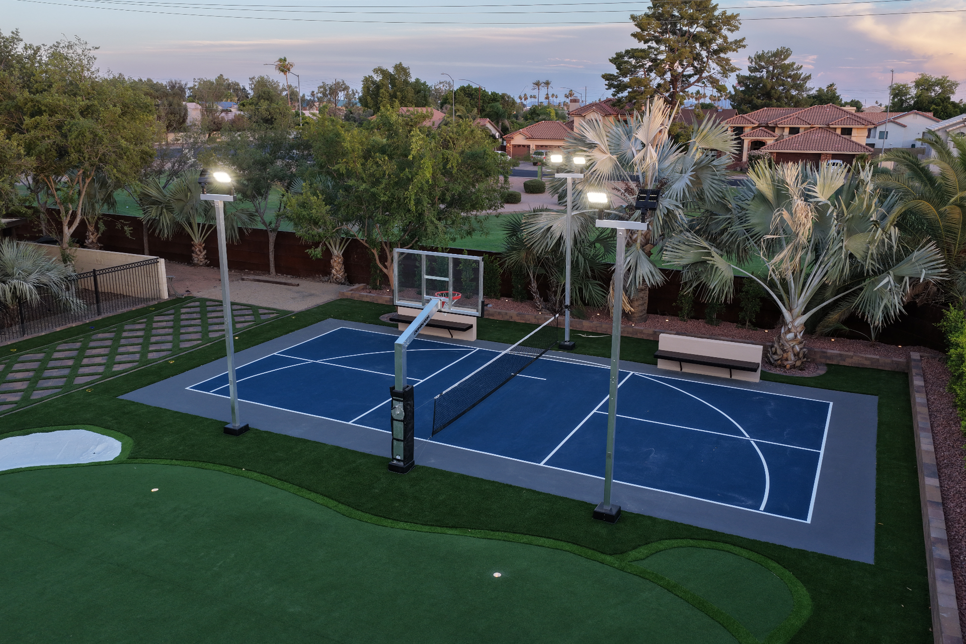 Blue multi-sport court with basketball hoop, net and LED light poles at dusk
