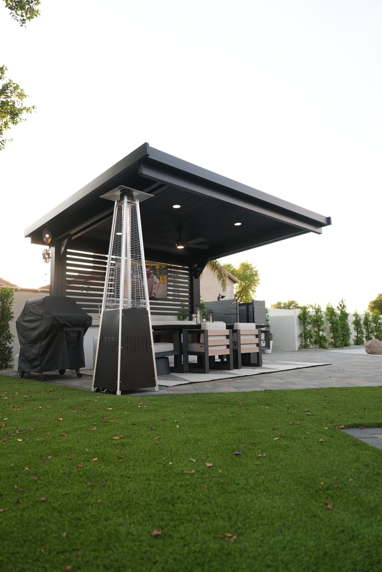Black pavilion with heater and lounge seating beside a turf lawn at dusk